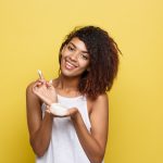 Beauty Concept – Beautiful young African American woman happy using skin care cream. Yellow studio background. Copy Space.